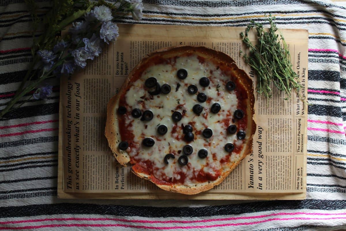 Sourdough discard pizza crust made into a home sourdough discard pizza with toppings and cheese sitting on a parchment lined cutting board, on a tea towel with flower, herbs and a pizza cutter around it. 