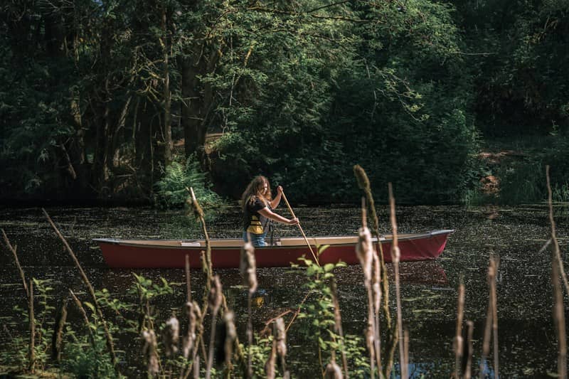 A woman paddling a red canoe on a dark pond surrounded by trees