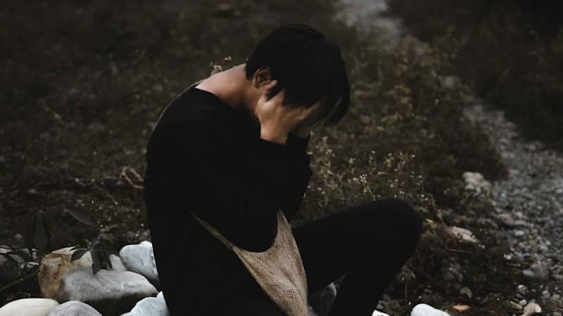 person in black shirt and gray pants sitting on ground  with their head in their hands emotionally processing. Our bodies hold on to trauma, join us for Book Club.