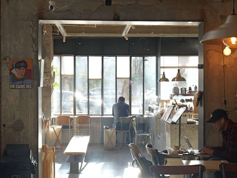 Man working on laptop in a sunlit cafe. He's not taking action but he's working.