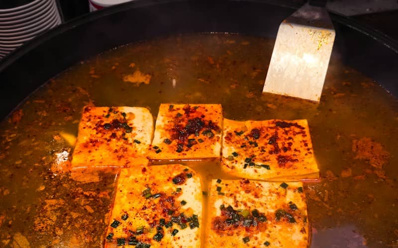 Four squares of tofu simmering in a spicy broth with green onions