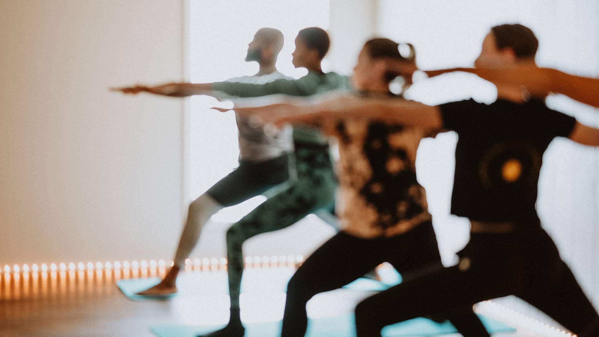 A group of people practicing yoga indoors, performing a standing pose with their arms extended. They are on yoga mats in a well-lit room. The image is slightly blurred, giving a soft focus to the scene.