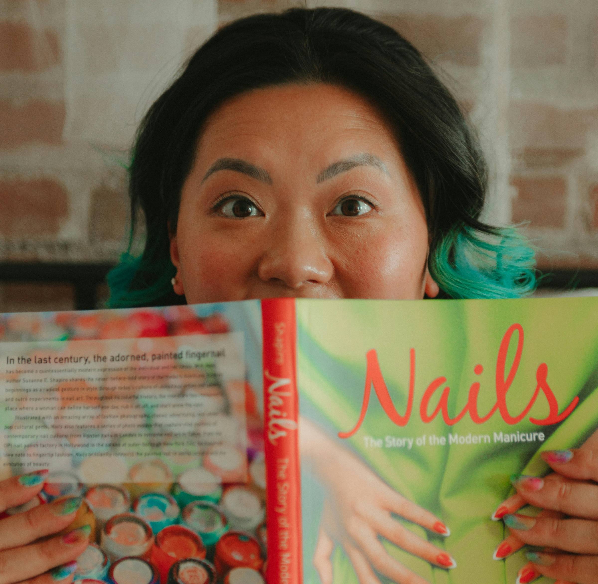 Mary is smiling behind a book called "Nails". The book is obscuring the bottom half of her face.