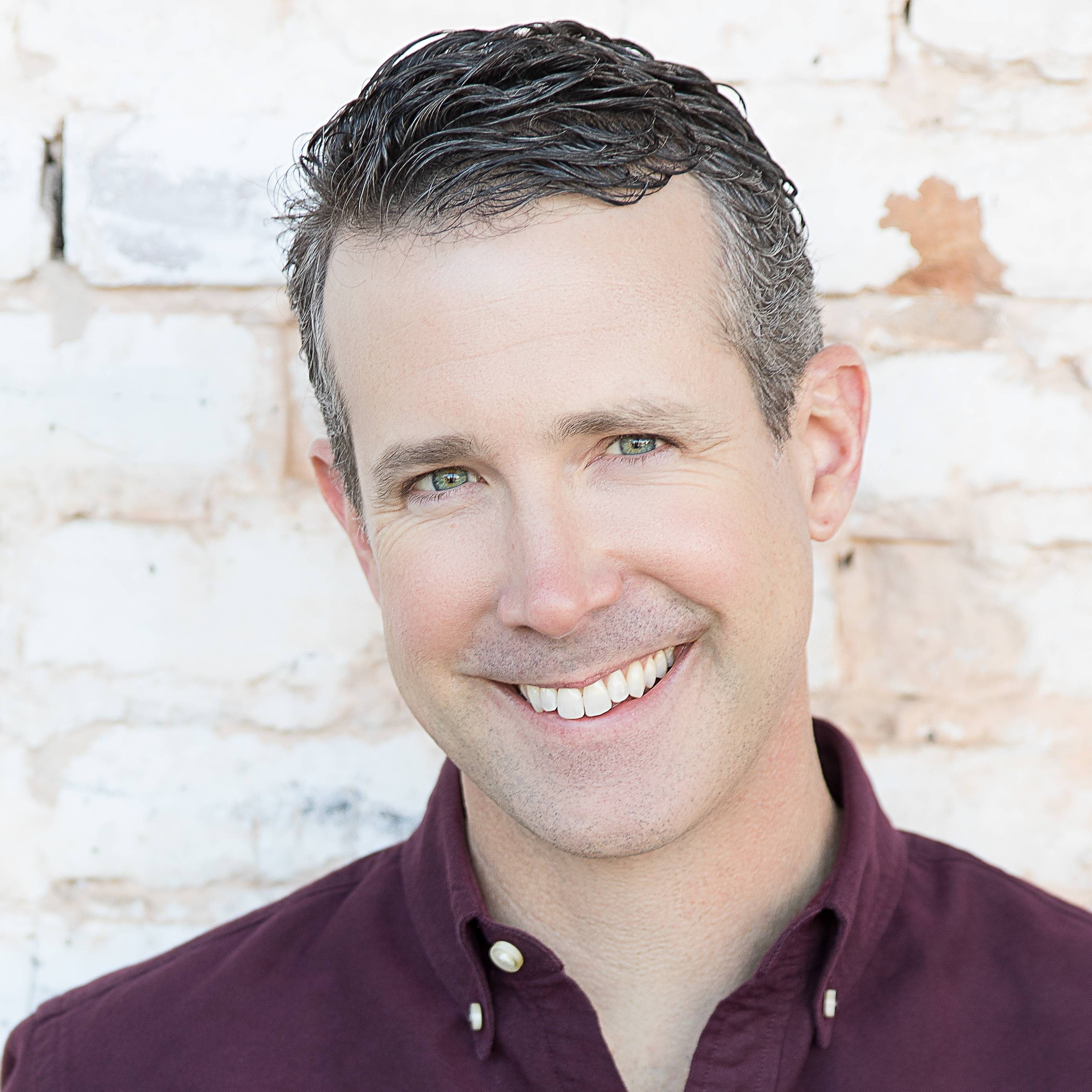 A person smiling at the camera, wearing a dark maroon button-up shirt. The background is an off-white, textured brick wall. The individual has short, dark hair and green eyes, conveying a friendly and approachable demeanor.