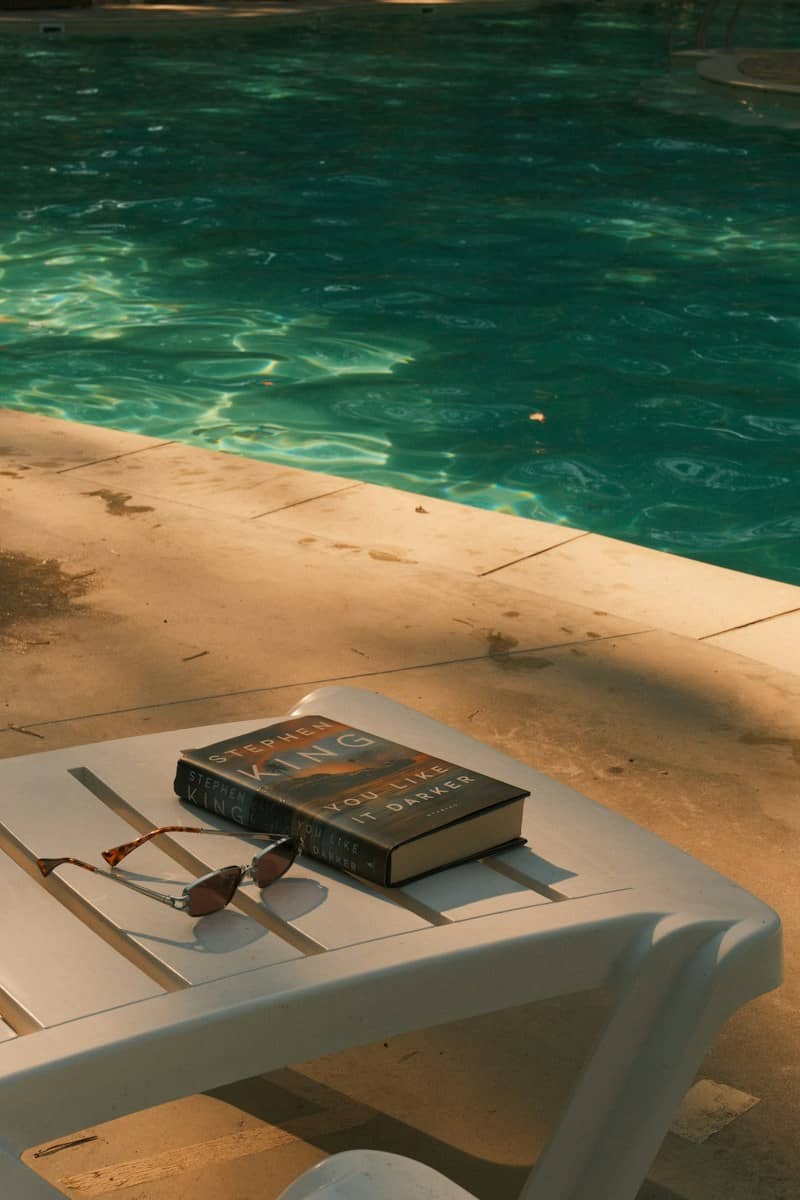 A book and sunglasses on a white plastic lounge chair by a pool