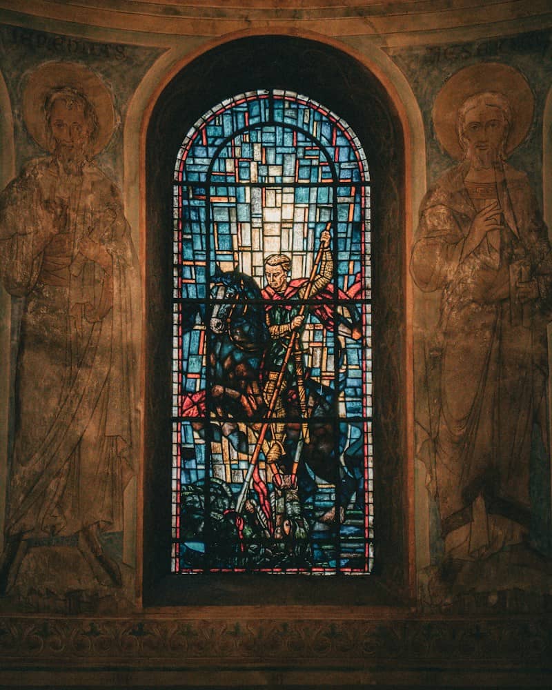 Stained glass window depicting a knight on horseback flanked by two painted figures