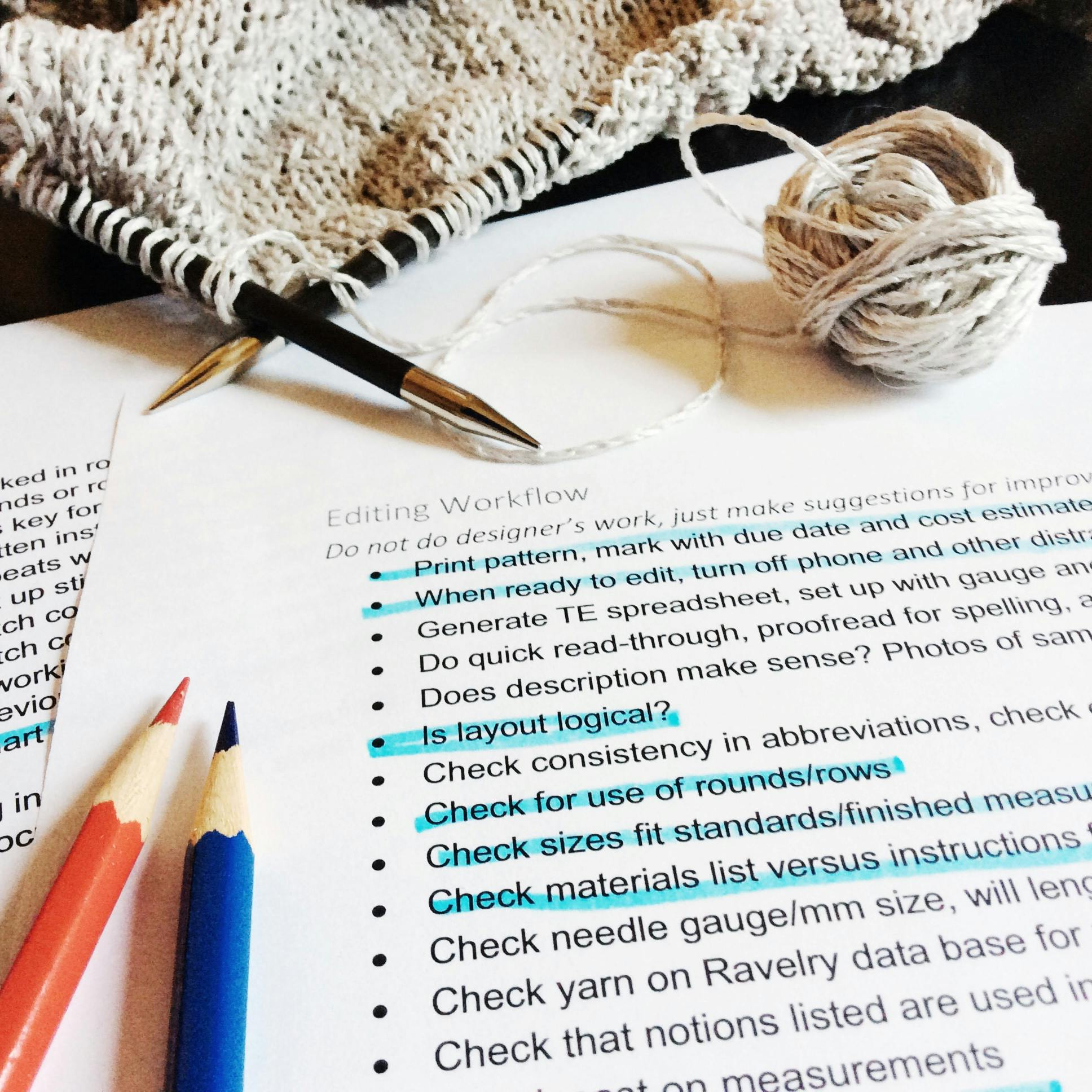 A red and blue pencil sit next to a tech editing checklist as well as a knitting project and grey yarn