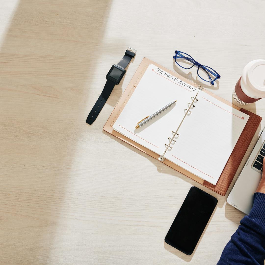 A desk planner sits on a wooden desk with glasses and a watch while a person works on their laptop