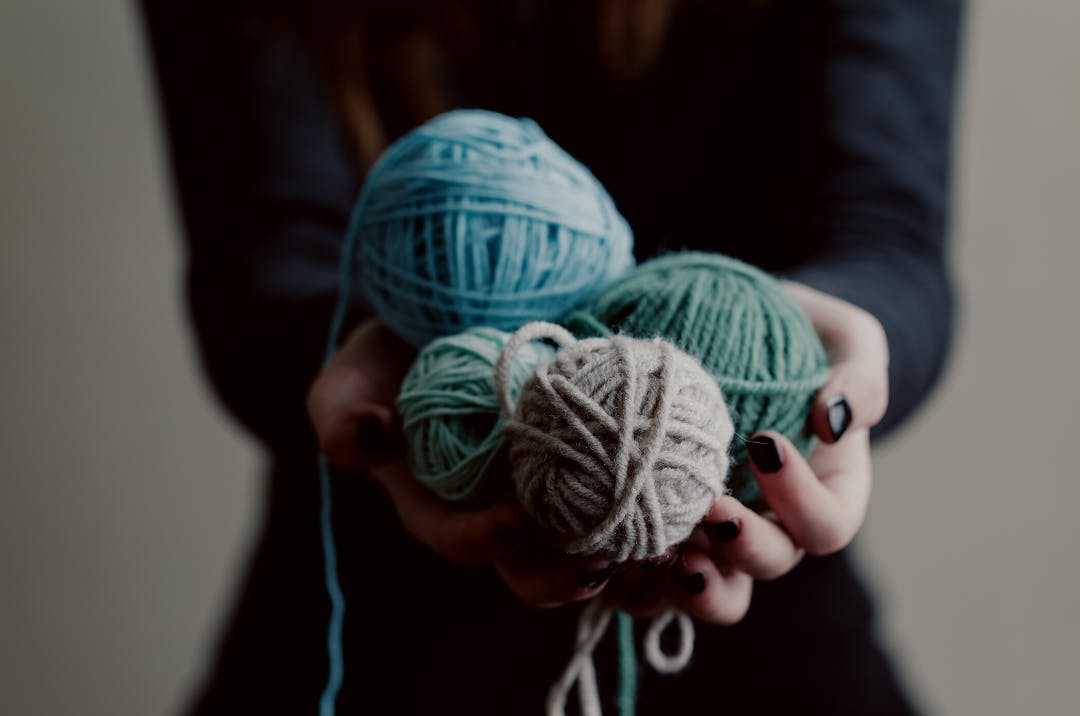 A woman with black nail polish on holds 4 balls of yarn in shades of blue, green, and grey.