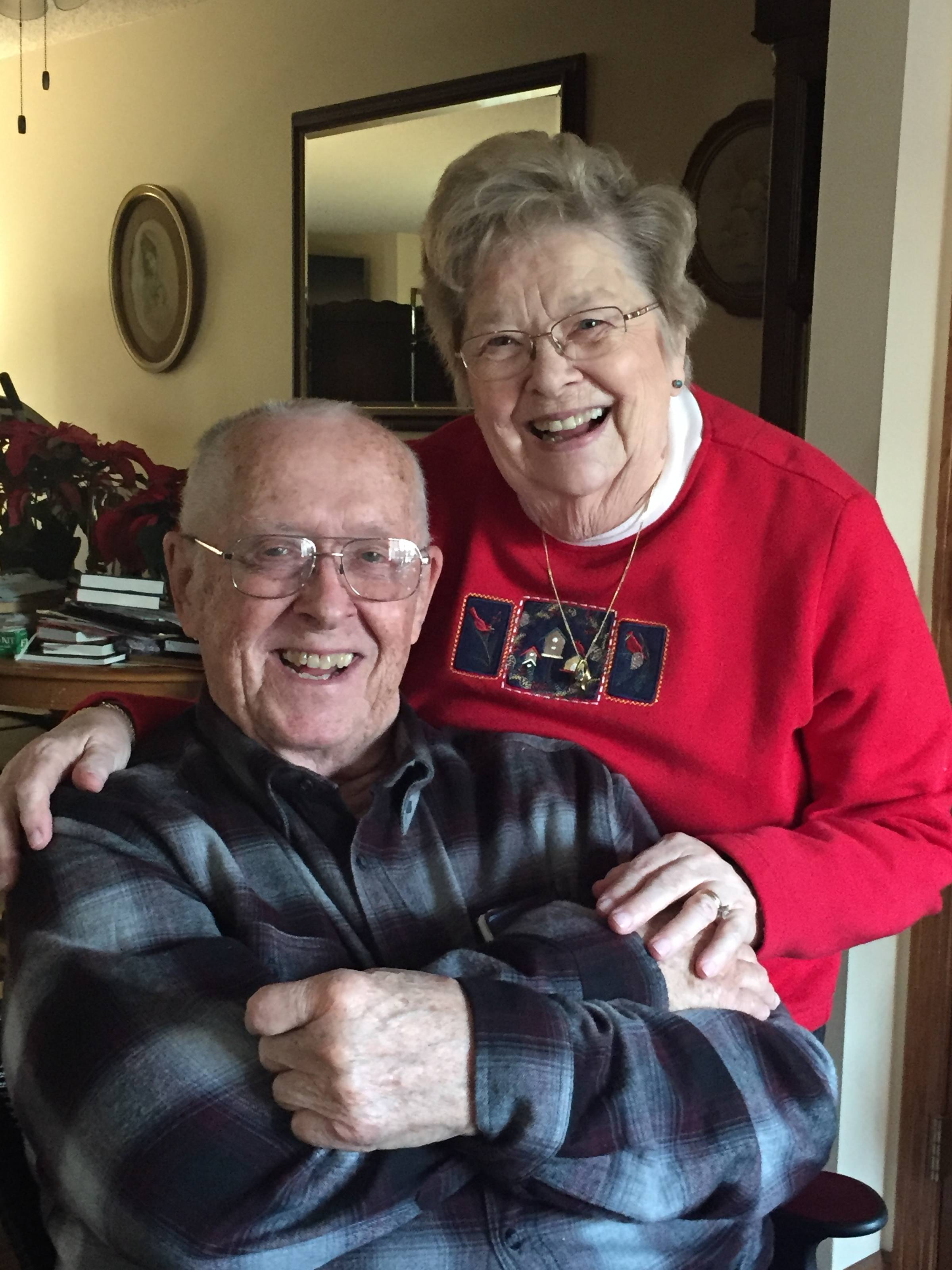 An elderly couple smiling together in a cozy indoor setting, the woman standing behind the seated man with her arm around him.