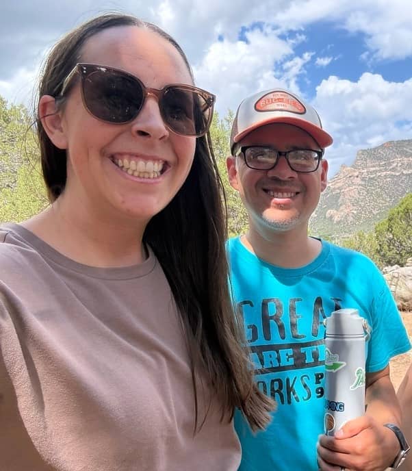 Smiling couple standing outdoors together in a mountain landscape beneath a bright blue sky with scattered clouds.