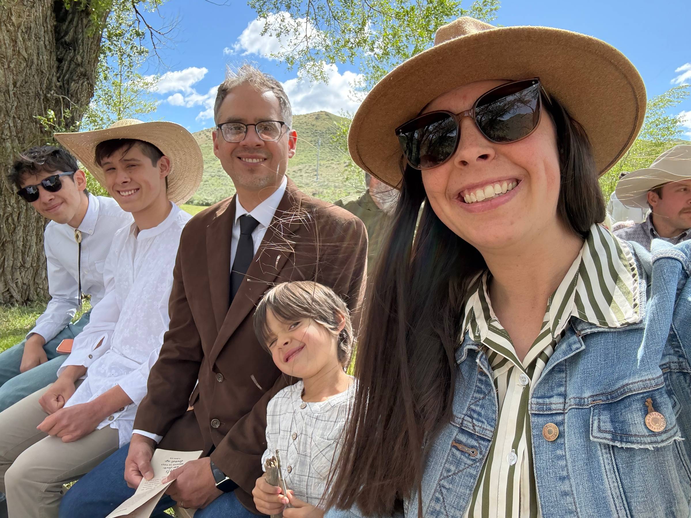 A family selfie at an outdoor western-themed wedding, featuring a smiling mom in a tan hat and denim jacket alongside her husband and three children seated together under a large tree. The family is dressed in western-inspired attire, with sunny skies, gr