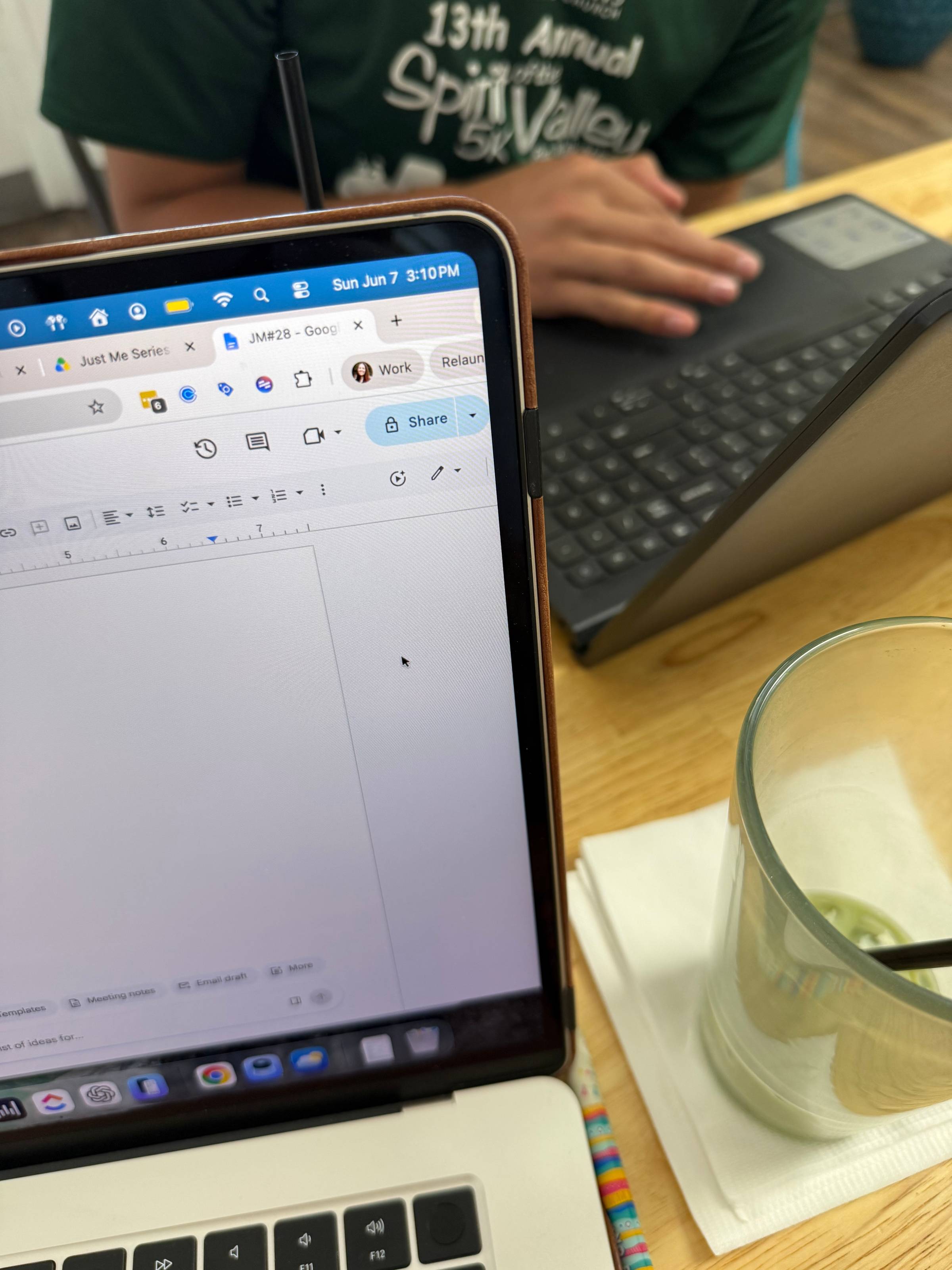 A laptop displaying a blank draft of JM#28 sits on a table at Kiln coffee bar, with a nearly empty drink beside it and a teenage boy working on a laptop in the background. The photo was taken on the one-year anniversary of his Type 1 Diabetes diagnosis, a