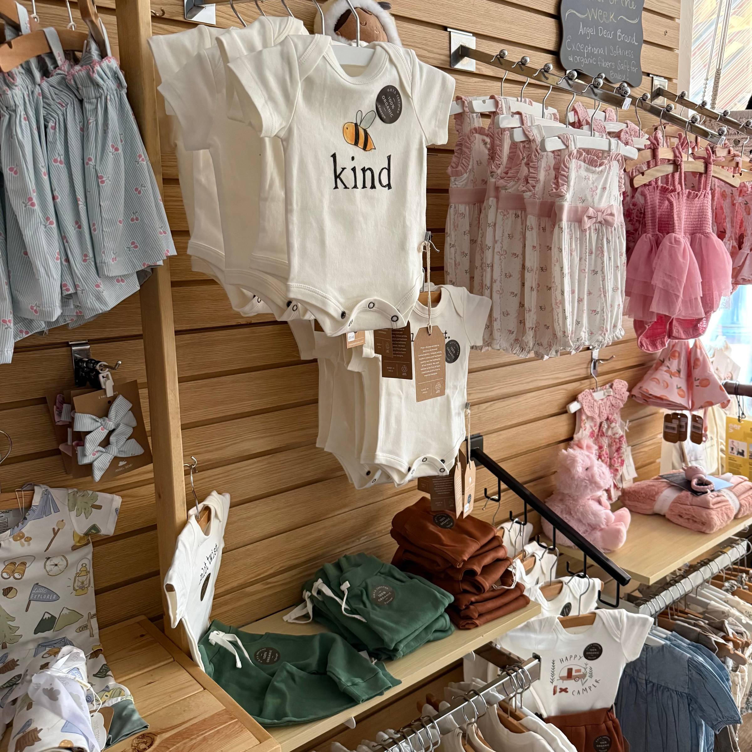 Display of organic baby clothing on wooden slat wall, featuring a white bee “kind” onesie, pink floral rompers, pastel dresses, bows, and folded baby joggers neatly arranged on shelves and hangers.