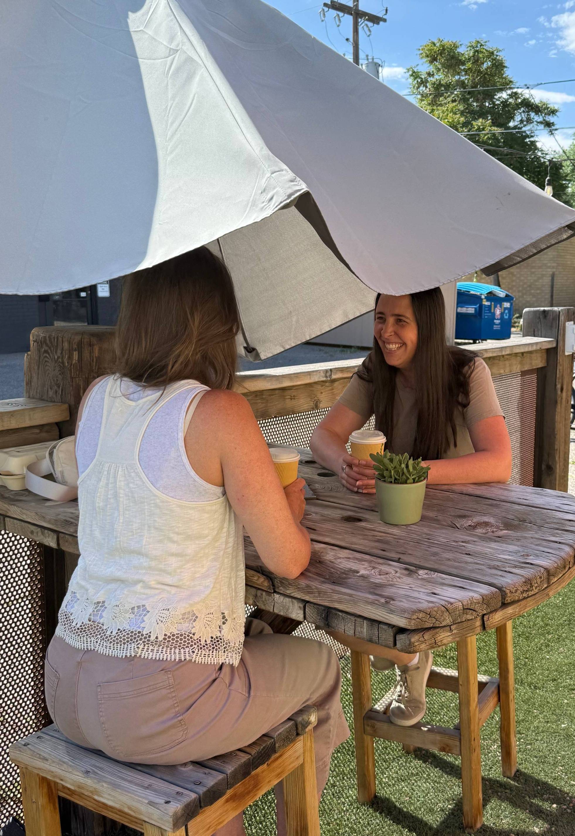 Two women sit across from each other at a rustic wooden patio table outside on a sunny morning, holding coffee cups and smiling mid-conversation. A small succulent sits between them on the table beneath a large umbrella, with downtown buildings, trees, an