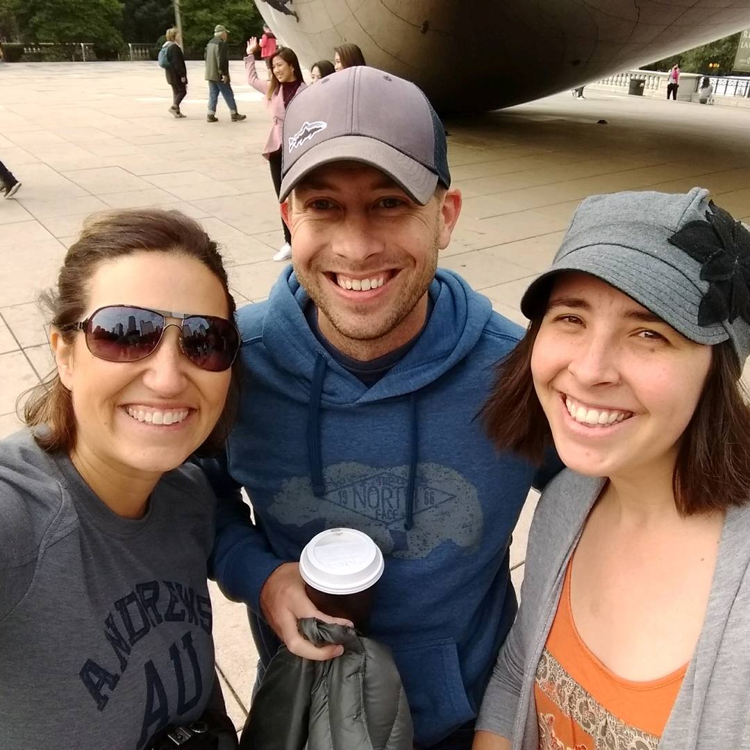 Three friends smile for a selfie outdoors near the reflective “Bean” sculpture in Chicago. One holds a coffee cup, and they’re all dressed casually in sweatshirts and jackets.