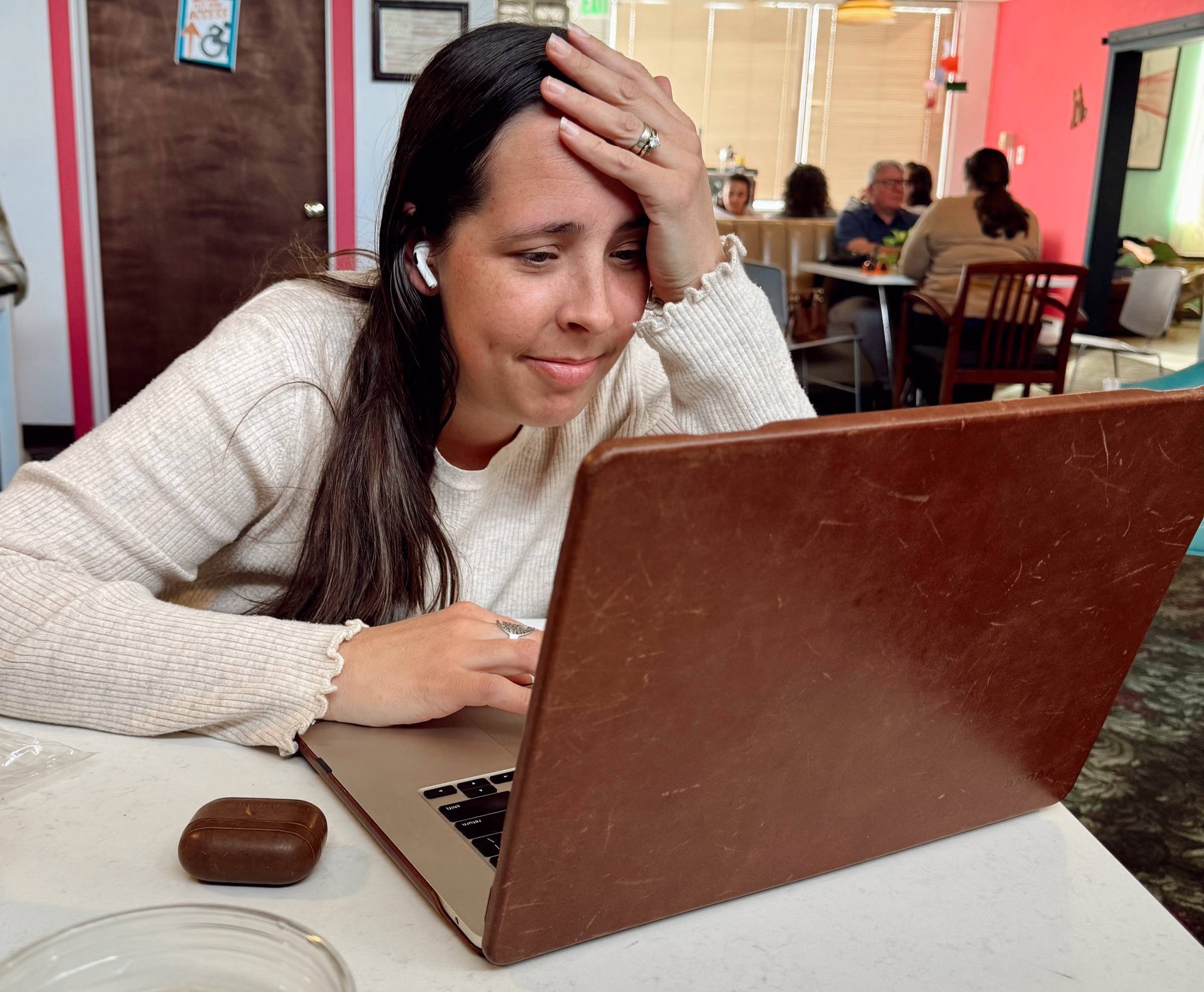 Megan sits at a table in a coffee shop, hand on her forehead, looking emotional as she reads something on her laptop. She’s wearing AirPods and a light knit sweater, with a glass of water and her earbuds case on the table. The moment looks tender and over