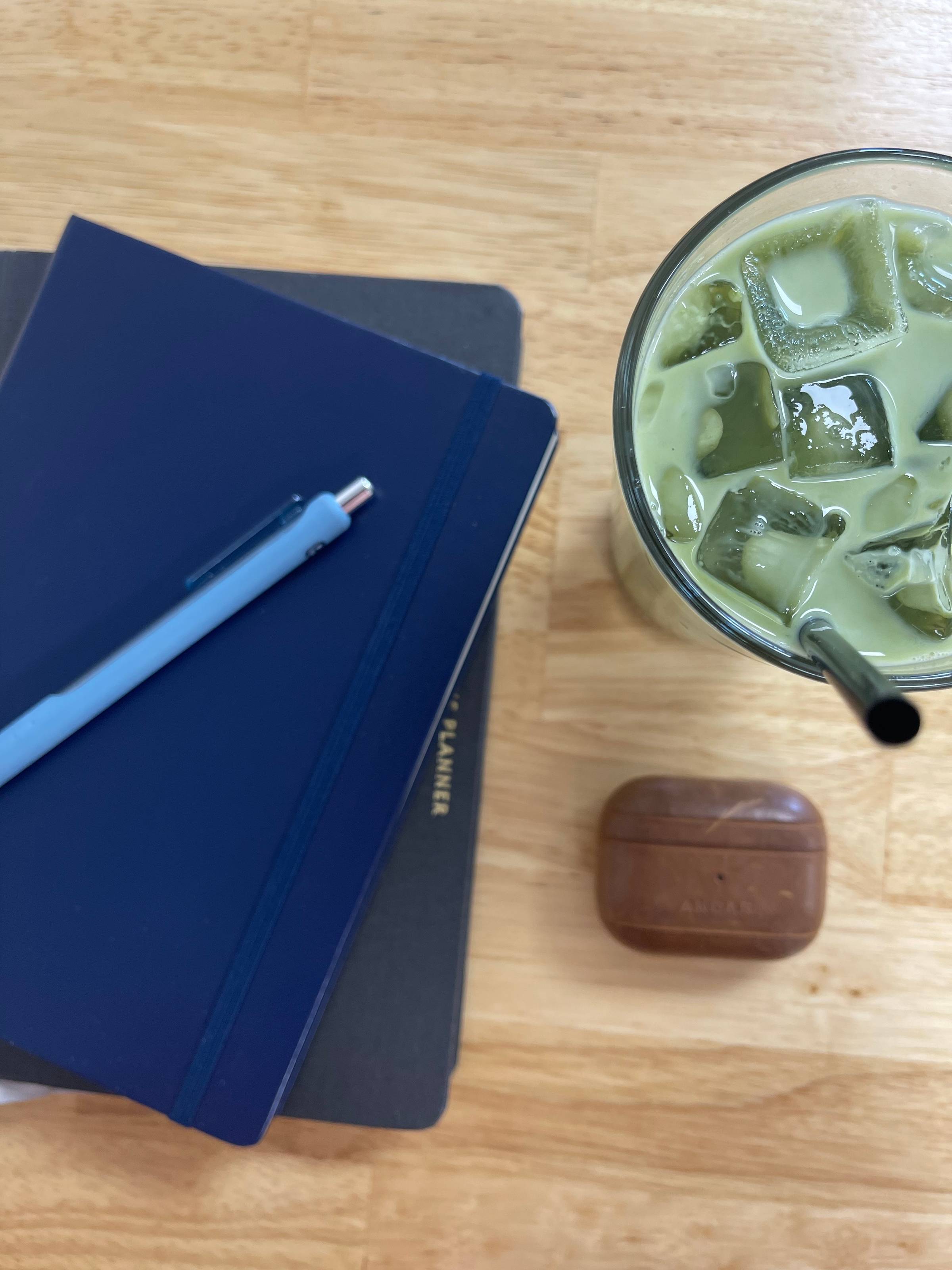 A navy blue journal and light blue pen resting on a wooden table beside an iced matcha latte and a brown AirPods case — a quiet little snapshot of writing, reflection, and slowing down.