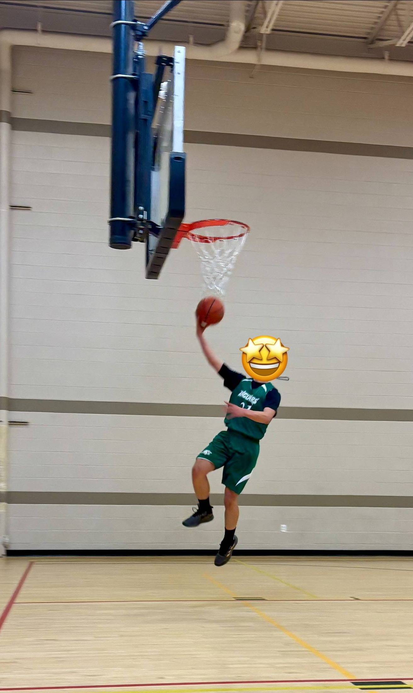 A boy in a green basketball jersey jumps toward the hoop for a layup during a game in a gymnasium.