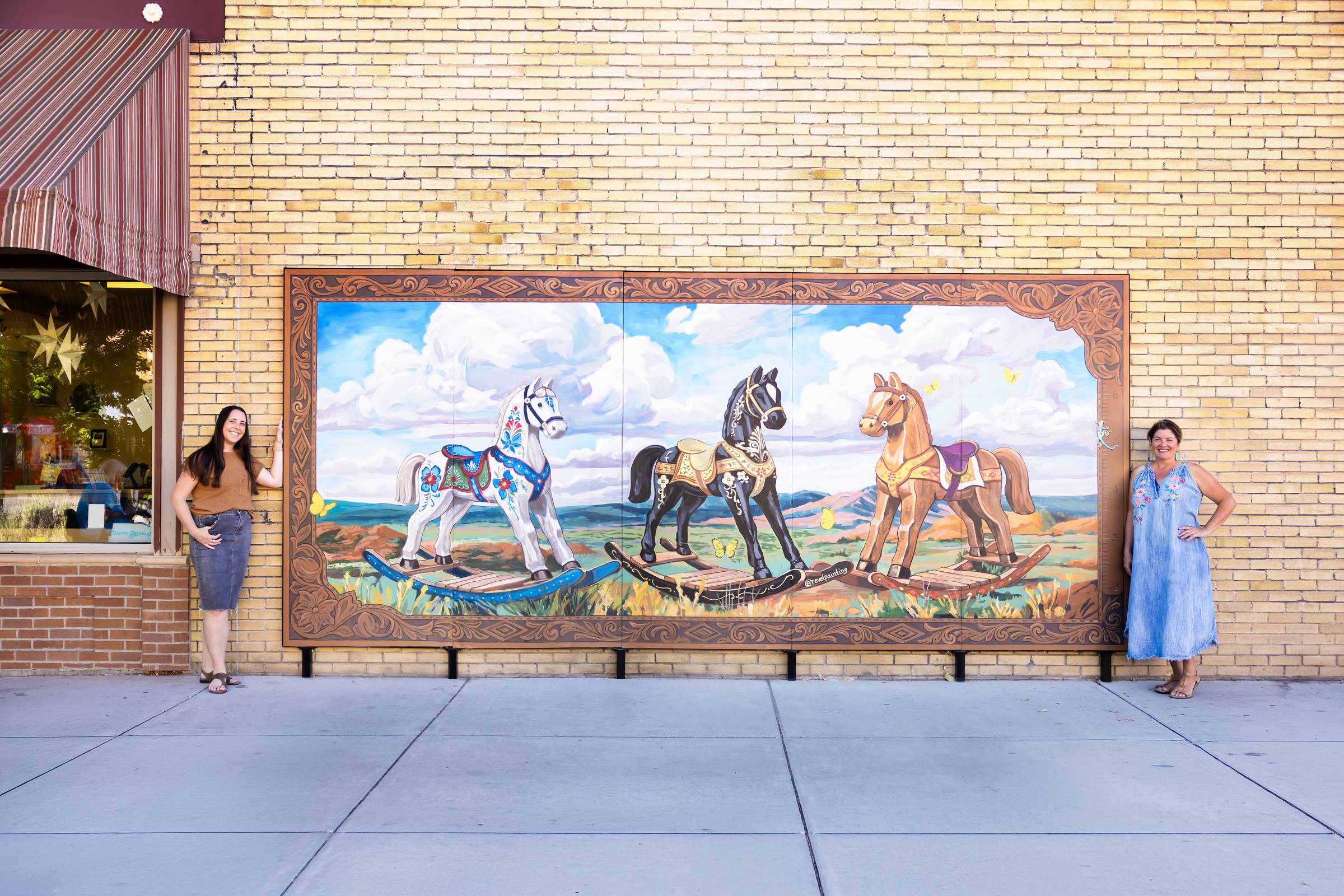 Megan and muralist Hilary standing together in front of the completed Colorado Baby mural, featuring three rocking horses against a colorful Western Slope landscape.