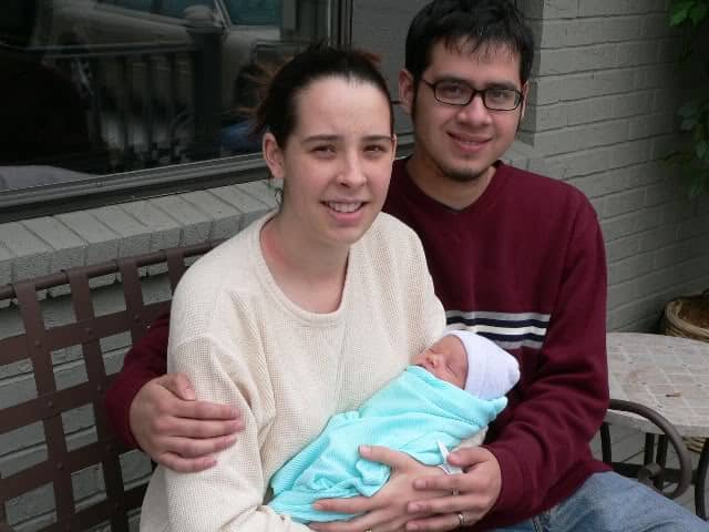 A young mother and father sit together on a bench, the mother cradling their newborn baby swaddled in a mint-green blanket while both parents look toward the camera.