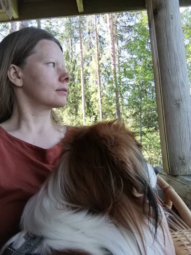 Miten maadoittua luonnossa. Nainen ja koira istuvat erämökin kuistilla. Suomalainen havumetsä siintää takana.