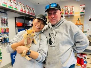 A couple with sweat shirts and baseball caps holding a red tabby kitten and smiling at the camera