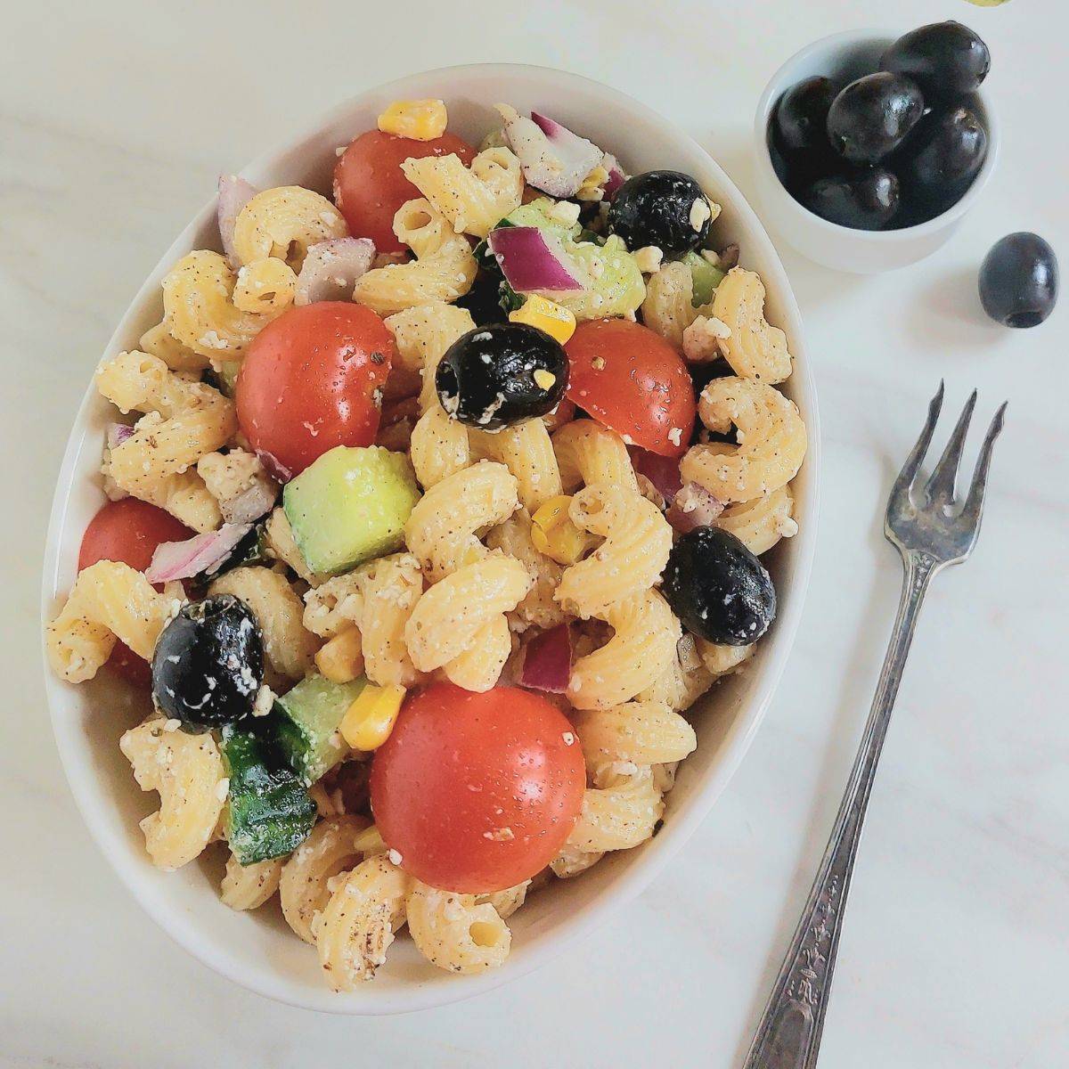 white oval shaped bowl filled with greek vegetable pasta salad placed on marble along with a fork and a bowl of black olives.