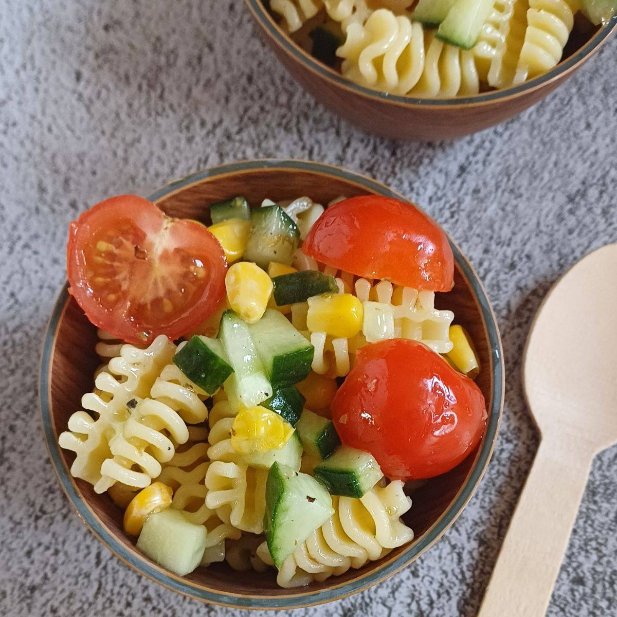 brown bowl of vegetable pasta salad placed on grey backdrop along with a wooden spoon.