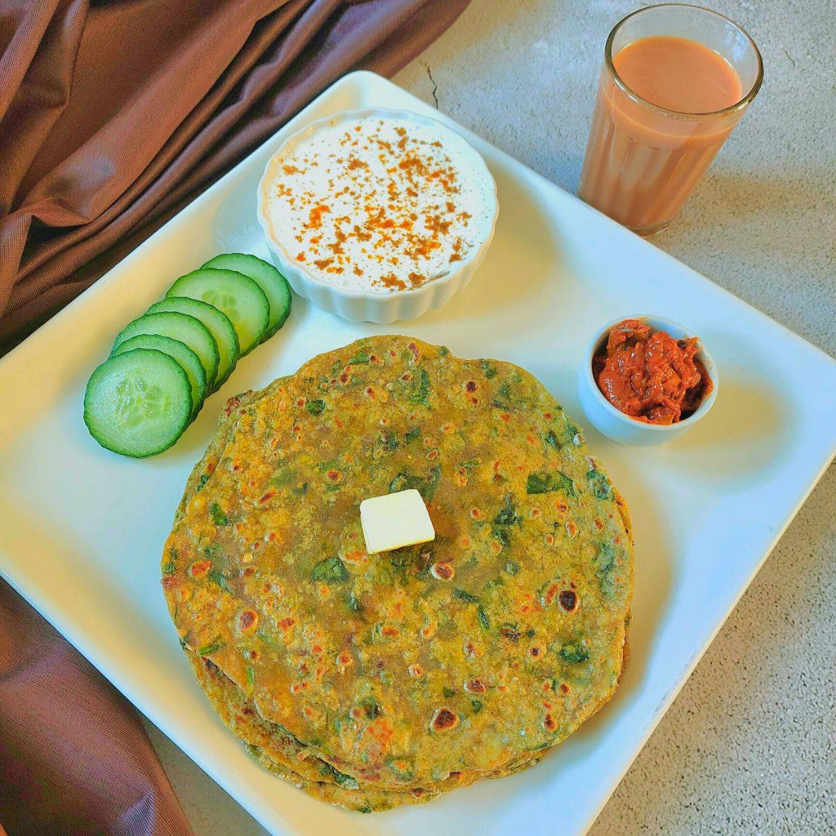 stacked aloo palak parathas on a square white plate along with cucumber slices, bowl of yogurt, pickle and a glass of tea placed behind it. to i