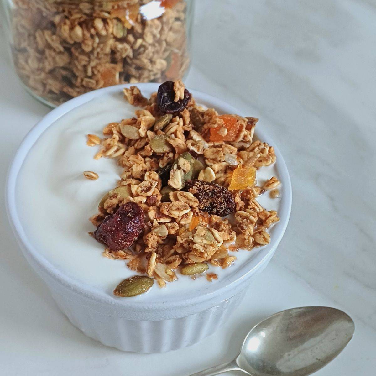 white bowl of yogurt topped with crunchy oil free granola placed on marble with spoon next to it and a jar of granola behind it.