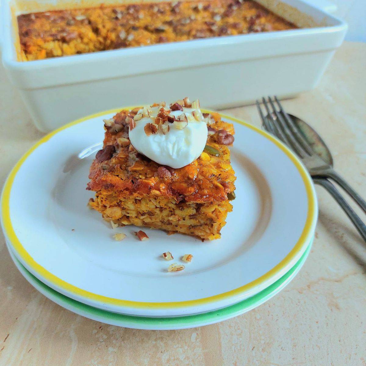 slice of baked pumpkin oatmeal on a white plate placed on granite along with cutlery and a tray of it behind it.