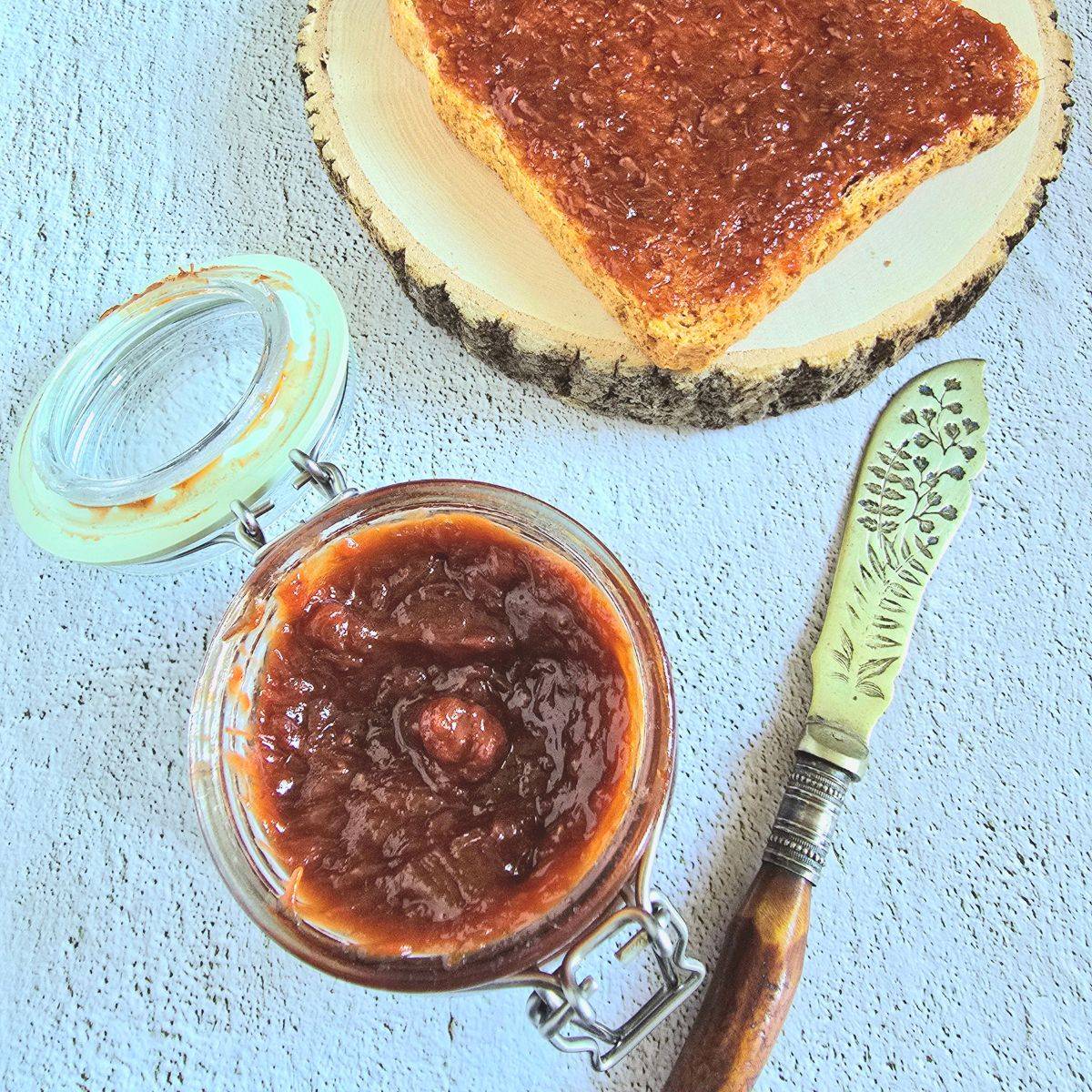 glass jar of strawberry rhubarb jam placed on blue backdrop along with a knife and a toast on wooden block. 