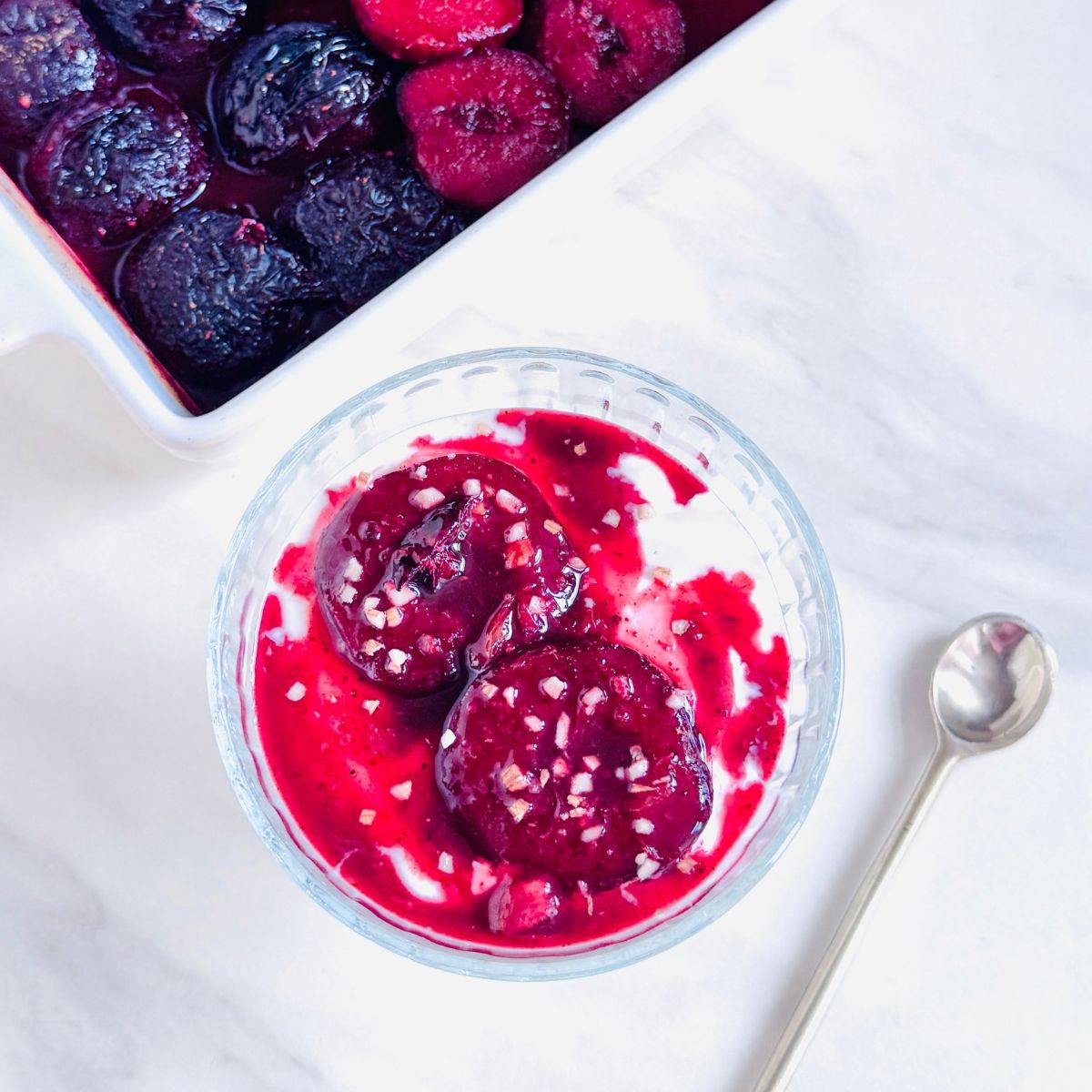 front image of bowl of yogurt topped with baked plums placed on marble along with a spoon.