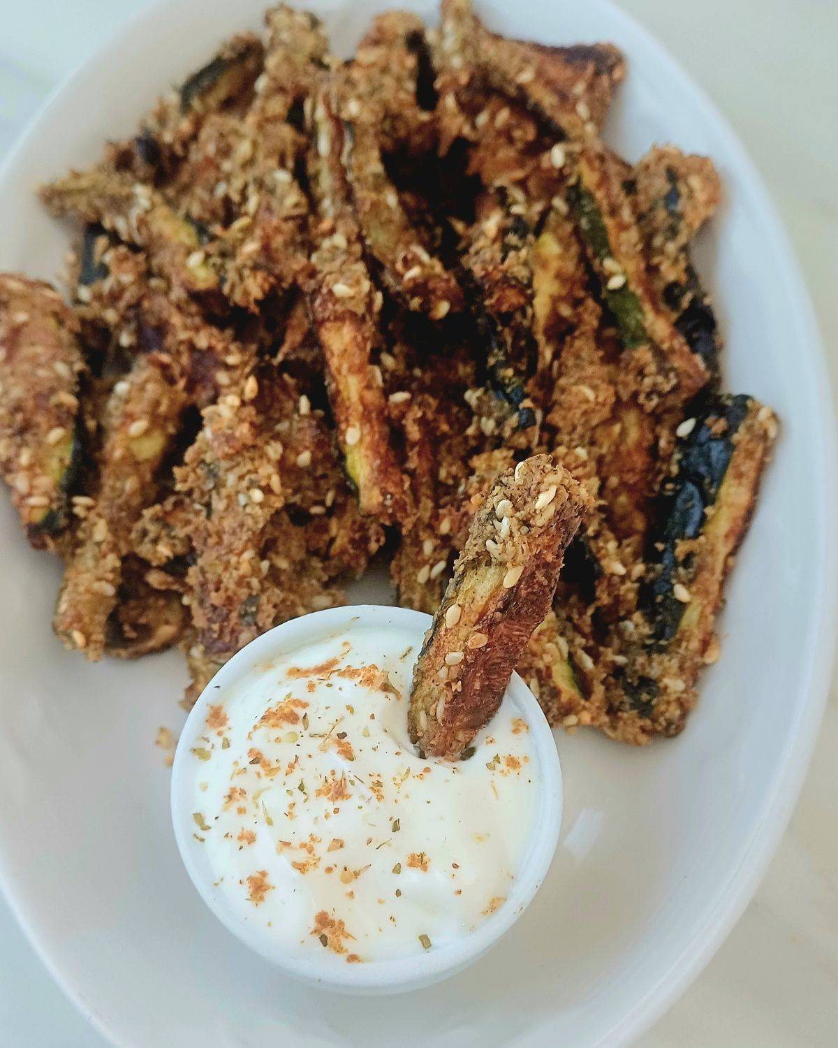 crunchy baked zucchini sticks on white plate along with a bowl of yogurt dip. 
