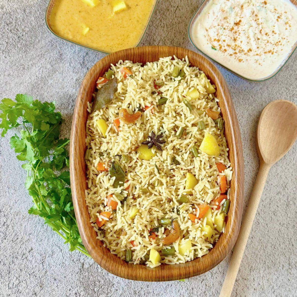 brown container of vegetable coconut milk pulao placed on grey background along with a bunch of coriander, wooden spoon, bowls of yogurt and curry