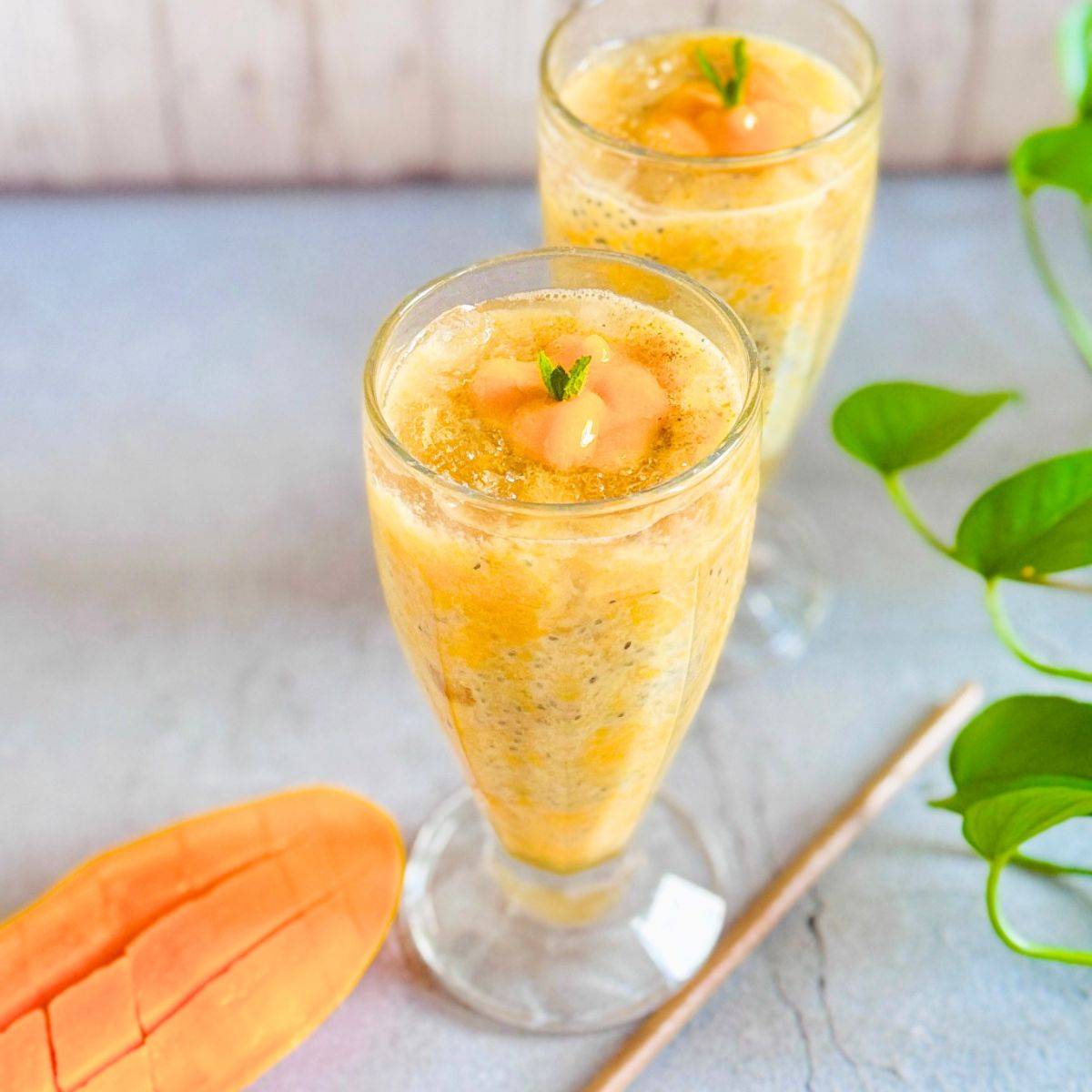 two glasses of mango gond katira sabja drink placed on a granite along with half cut mango and eco friendly coconut leaf straw.