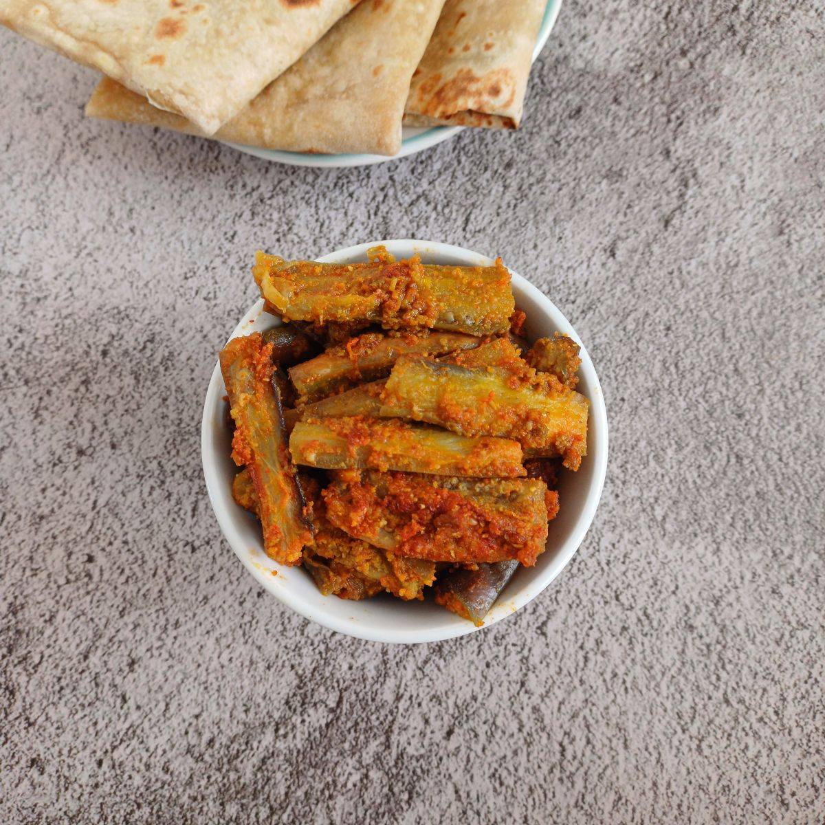 white bowl of spicy japanese eggplant dish placed on grey backdrop along with a plate of chapatis.