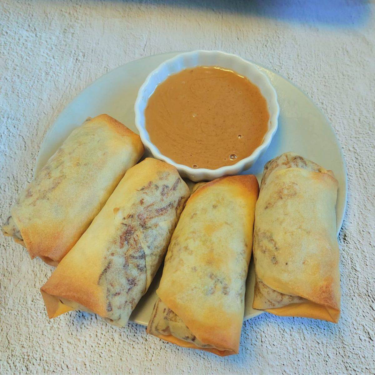 four baked vegetable spring rolls on a grey plate along with a bowl of peanut dipping sauce placed on black and white textured backdrop.