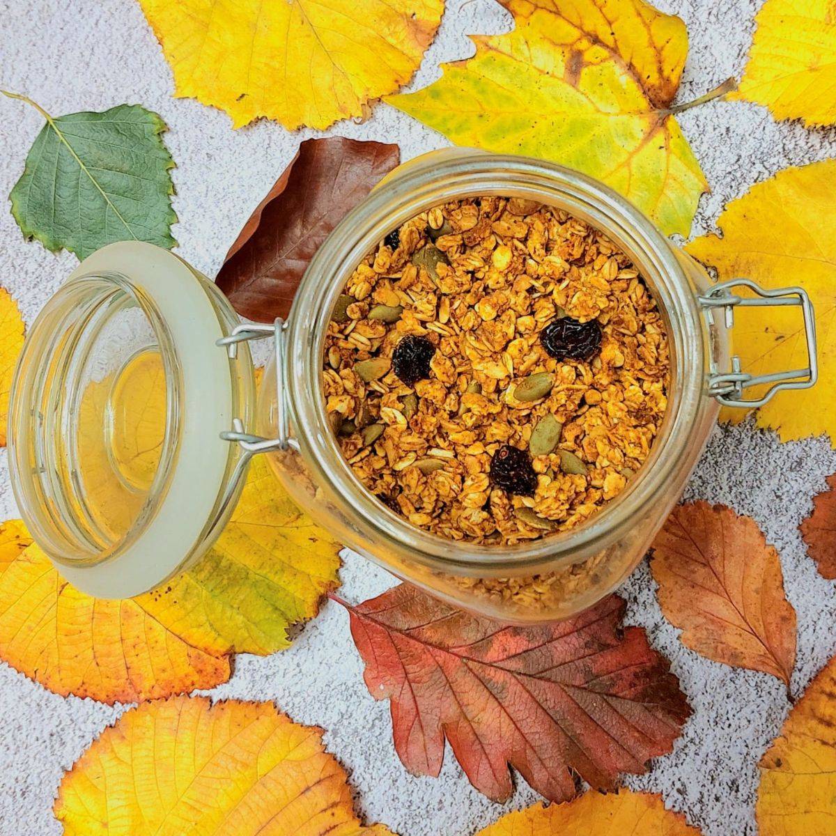 jar of pumpkin granola placed on grey backdrop with autumn leaves around it. 