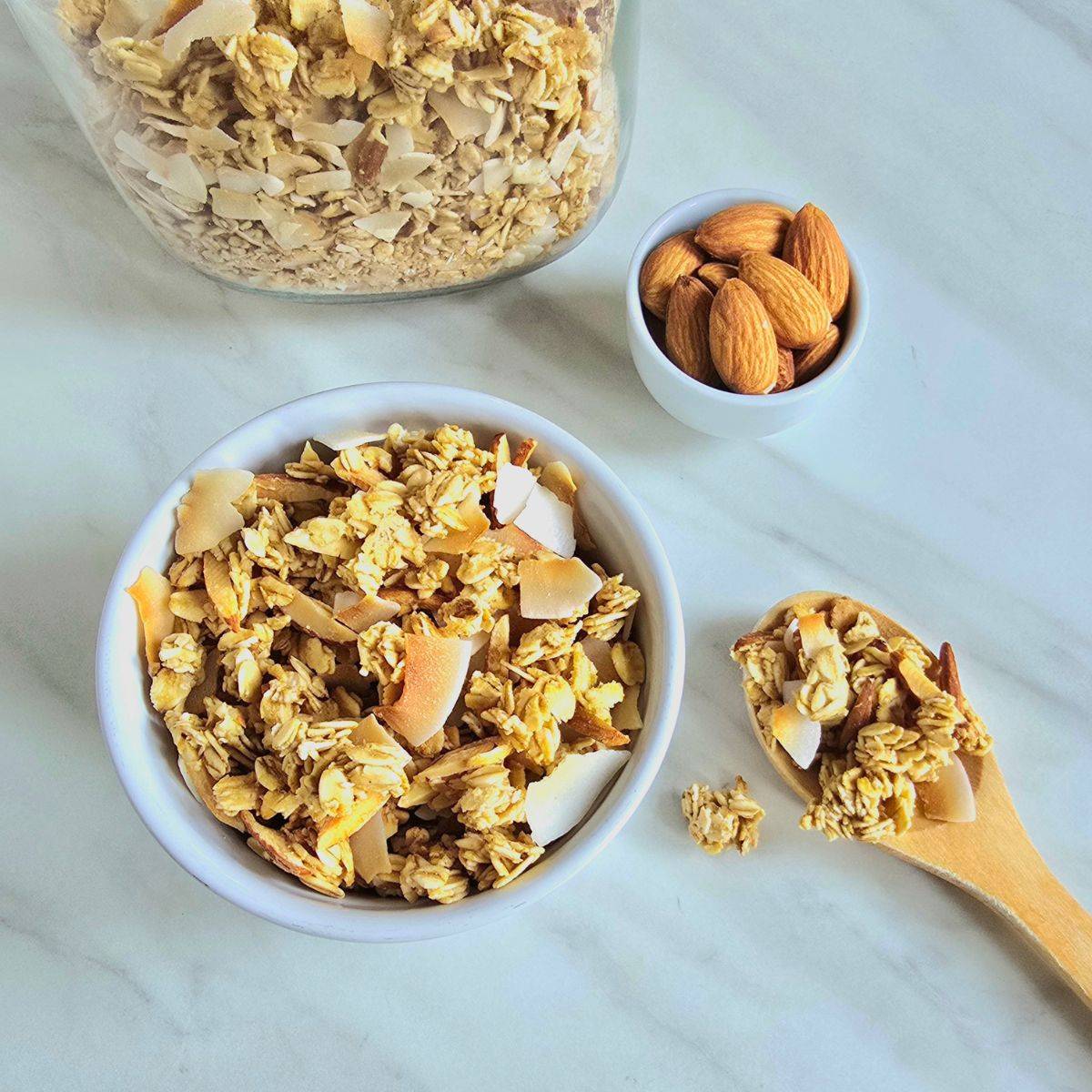 white bowl of coconut granola placed on marble along with a spoon of granola, pinch bowl of almonds and a glass jar of granola.