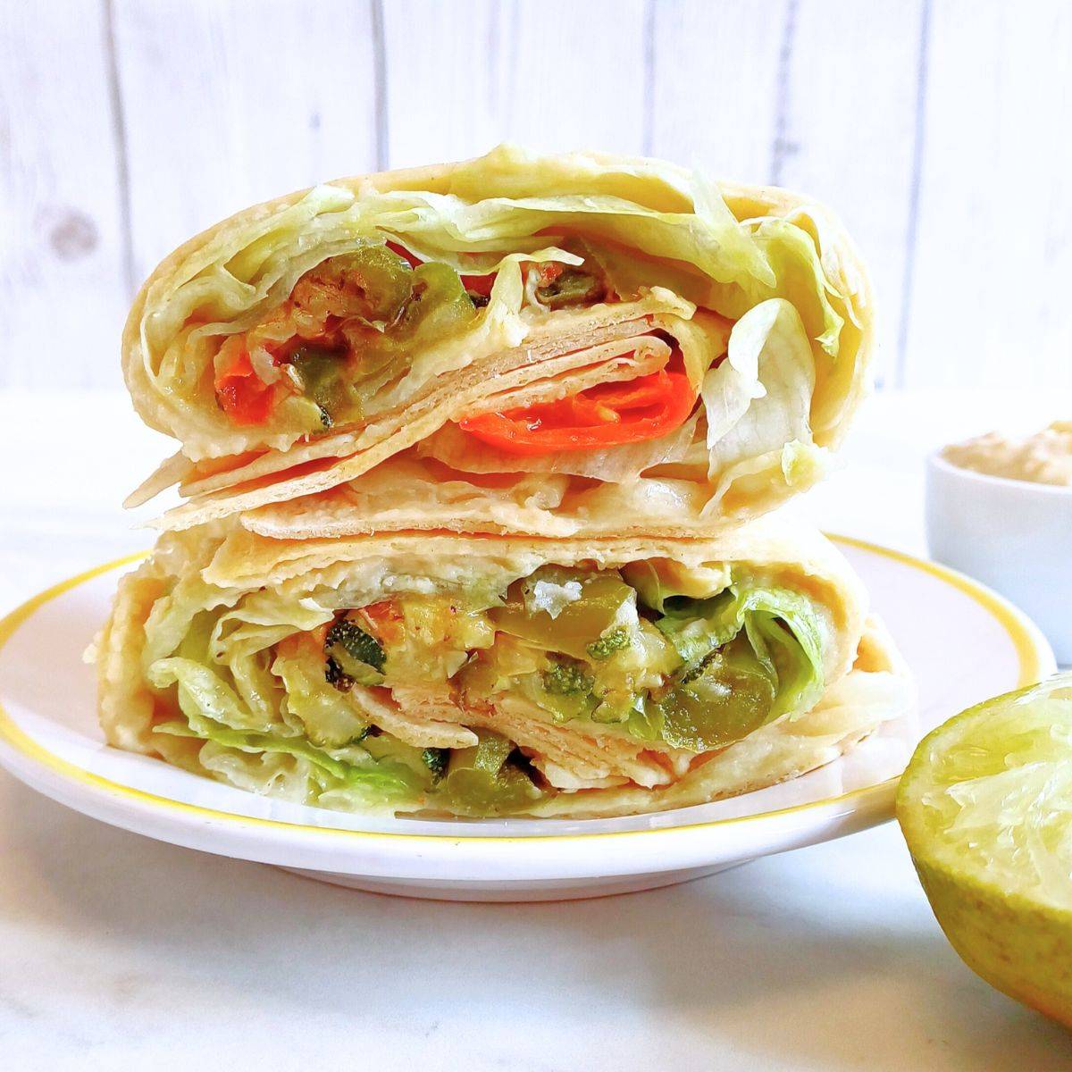 stacked roasted veggie hummus wraps placed on a white plate along with half lime and a pinch bowl of hummus next to it.