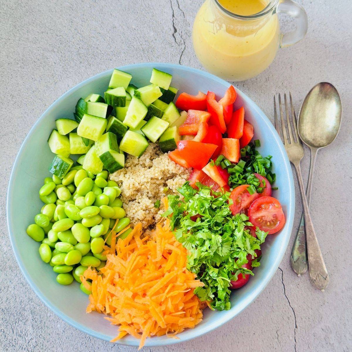 bowl bowl of quinoa edamame salad with peanut butter mango dressing placed on grey granite along with fork, spoon and a jug of dressing.