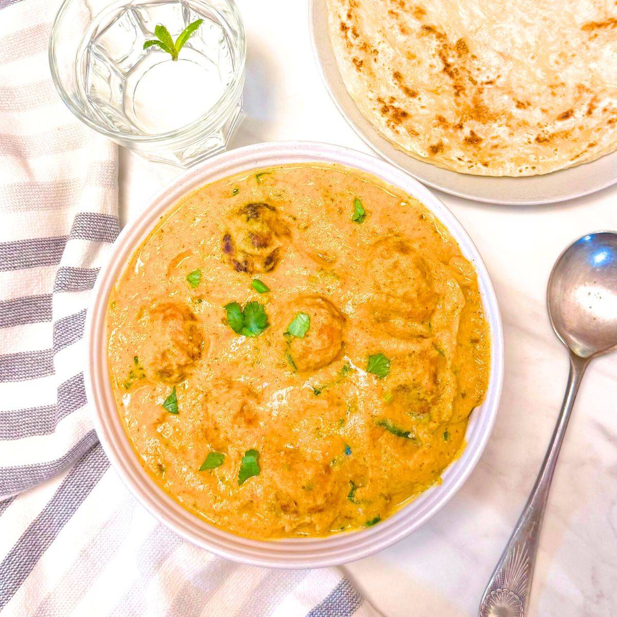 bowl filled with mixed vegetable kofta curry placed on marble along with a spoon, glass of water, parathas and a napkin