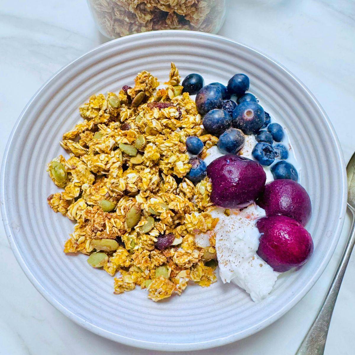 white bowl of oil free sweet potato granola, yogurt, cherries and blueberries placed on marble along with a spoon.