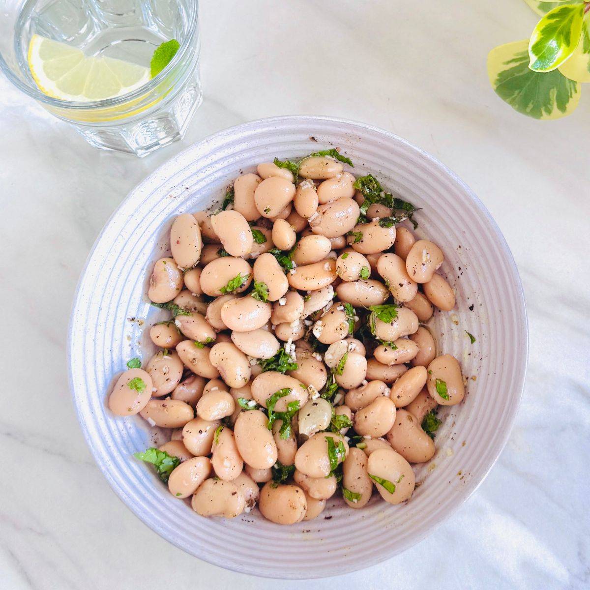 white bowl of butter bean salad with garlic herb dressing placed on marble along with a glass of water.