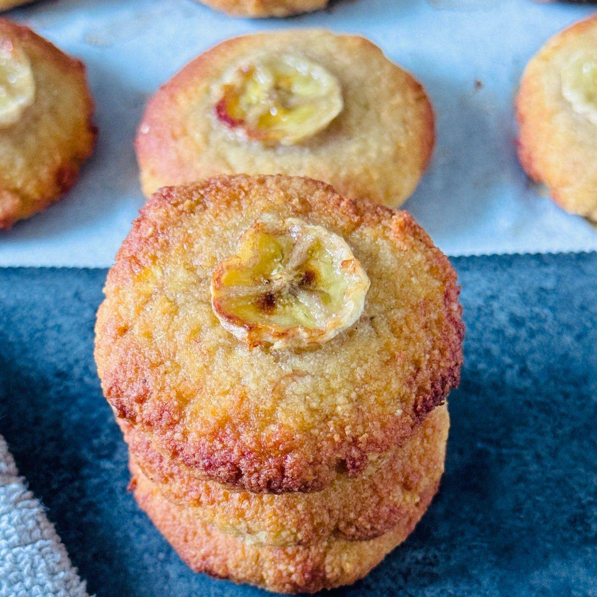 stack of banana almond cookies placed on a dark granite along with more cookies on baking sheet behind them.