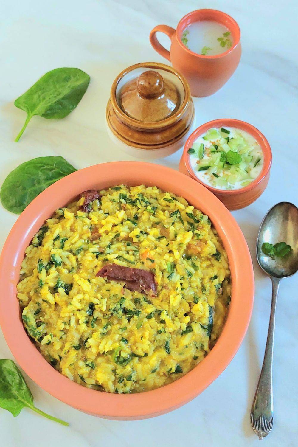 brown bowl of palak moong dal khichdi placed on marble along with a spoon, cup of raita, pickle bowl and a glass of lassi.