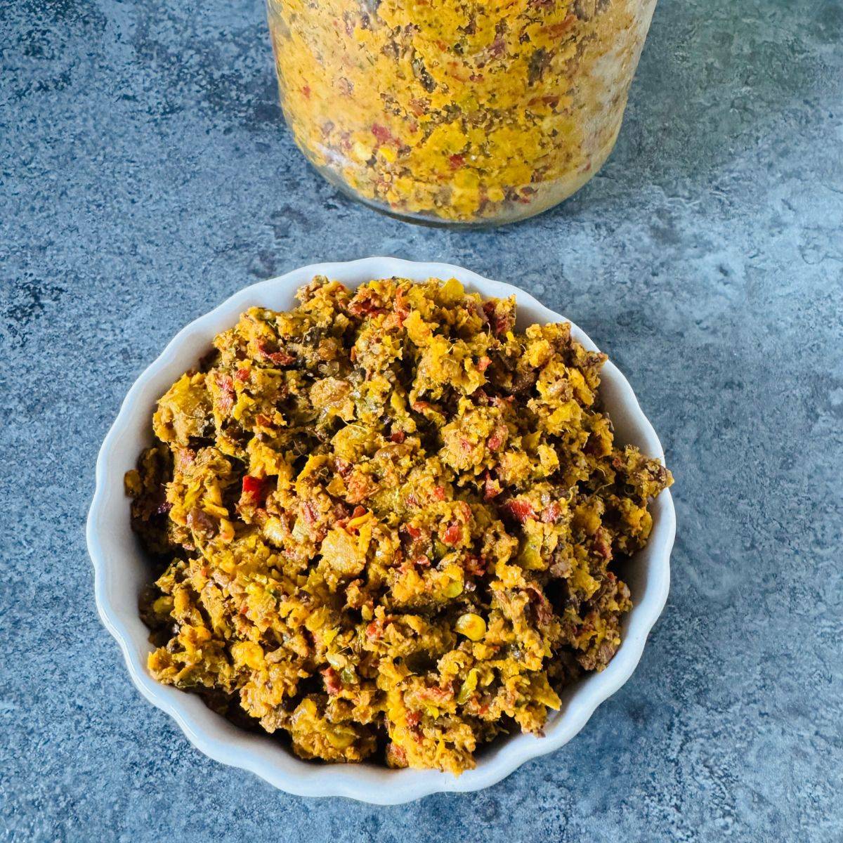 White bowl of nellikai thokku placed on a textured tile with a bottle of thokku behind it. 