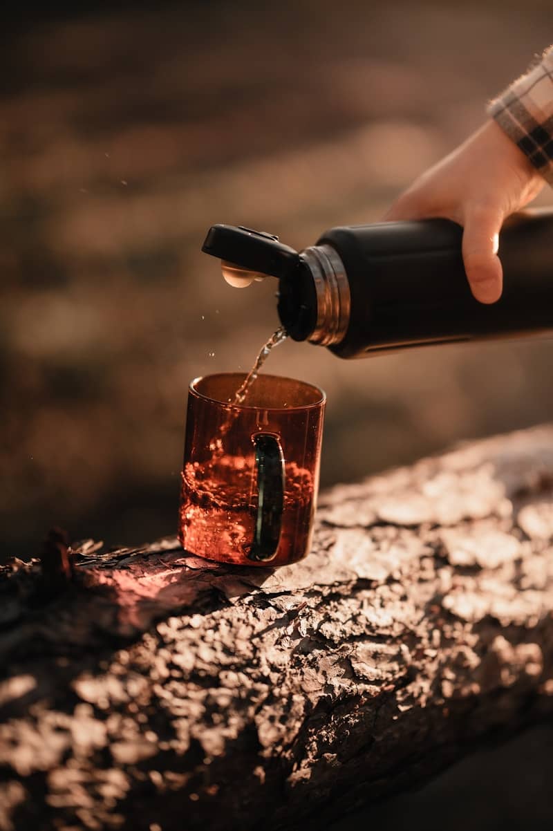 A person pouring liquid from a black thermos into a glass mug on a log